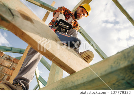 View from below, cutting plank by chainsaw. Construction worker is on the roof of a house View from below, cutting plank by chainsaw. Construction worker is on the roof of a house 137779323