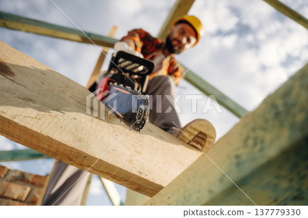 Doing the job, cutting the plank using chainsaw. Construction worker is on the roof of a house Doing the job, cutting the plank using chainsaw. Construction worker is on the roof of a house 137779330