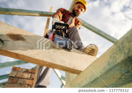 Serious facial expression, cutting the plank. Construction worker is on the roof of a house Serious facial expression, cutting the plank. Construction worker is on the roof of a house 137779332