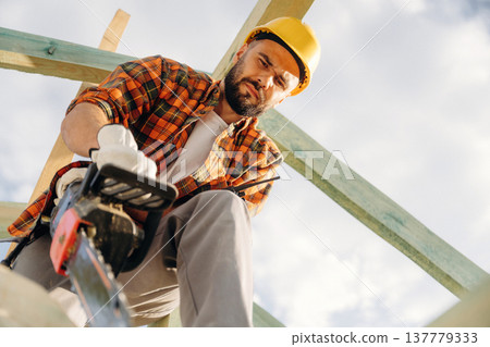 Serious facial expression, cutting the plank. Construction worker is on the roof of a house Serious facial expression, cutting the plank. Construction worker is on the roof of a house 137779333