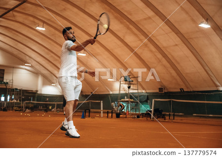 Side view, game in action. Man is playing tennis indoors in the hangar Side view, game in action. Man is playing tennis indoors in the hangar 137779574
