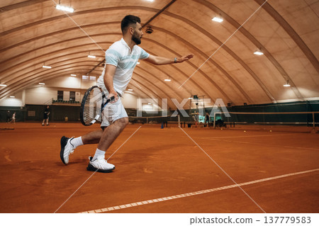 Running with racket, active time spending. Man is playing tennis indoors in the hangar 137779583