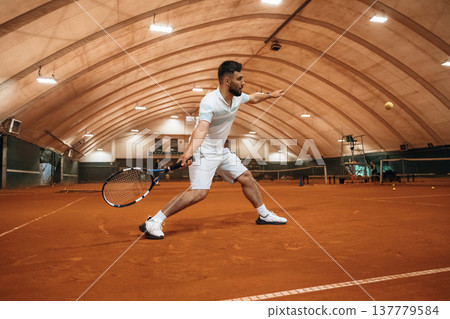 Running with racket, active time spending. Man is playing tennis indoors in the hangar 137779584