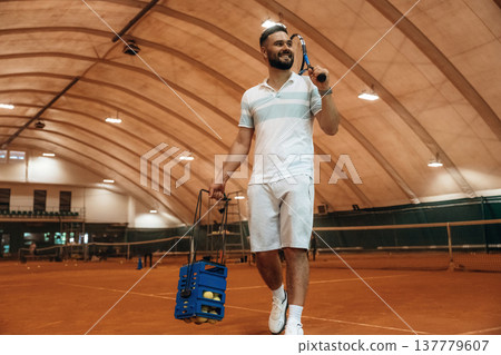 Walking forward, holding package with balls. Man is playing tennis indoors in the hangar 137779607