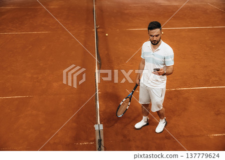Smartphone in hand, standing, taking a break, bored. Man is playing tennis indoors in the hangar 137779624
