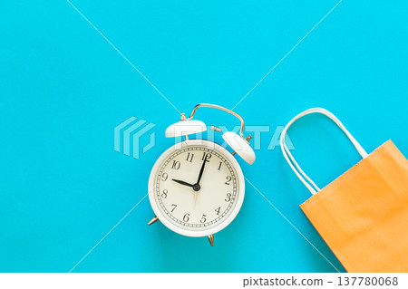 Paper bag and alarm clock on blue background, shopping time concept. Paper bag and alarm clock on blue background, shopping time concept. 137780068