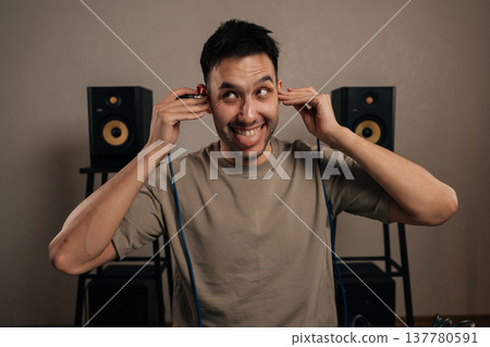 Portrait of cheerful male sound engineer making playful face listening through in ear monitors and adjusting audio in a professional recording studio, focused and cheerful during music production. 137780591