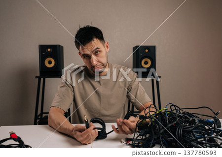 Portrait of frustrated man dealing with a massive pile of tangled electronic cables, facing a complex problem of wire management and technological clutter accumulation. Portrait of frustrated man dealing with a massive pile of tangled electronic cables, facing a complex problem of wire management and technological clutter accumulation. 137780593