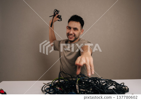 Vertical portrait of cheerful technician holding a large chaotic pile of tangled wires and cords, pointing finger directly forward, experiencing tech stress and cable management problems. Vertical portrait of cheerful technician holding a large chaotic pile of tangled wires and cords, pointing finger directly forward, experiencing tech stress and cable management problems. 137780602
