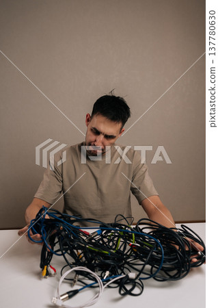 Vertical portrait of frustrated man dealing with a massive pile of tangled electronic cables, facing a complex problem of wire management and technological clutter accumulation. Vertical portrait of frustrated man dealing with a massive pile of tangled electronic cables, facing a complex problem of wire management and technological clutter accumulation. 137780630