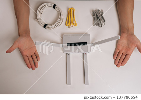Person arranging a white wireless internet router on a clean surface, surrounded by coiled ethernet cables and male hands, symbolizing network setup and connection. Person arranging a white wireless internet router on a clean surface, surrounded by coiled ethernet cables and male hands, symbolizing network setup and connection. 137780654
