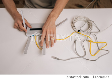 Close-up top view of technician hands connecting yellow ethernet cable into the port of a modern white wireless router with multiple antennas, symbolizing network setup and internet connectivity. Close-up top view of technician hands connecting yellow ethernet cable into the port of a modern white wireless router with multiple antennas, symbolizing network setup and internet connectivity. 137780663