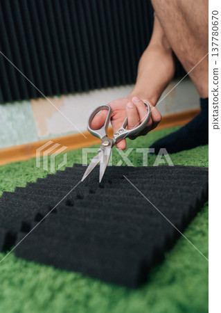 Person cutting a black acoustic foam panel on a green carpet with scissors, carefully customizing sound insulation material for a home recording studio or sound treatment project. Person cutting a black acoustic foam panel on a green carpet with scissors, carefully customizing sound insulation material for a home recording studio or sound treatment project. 137780670