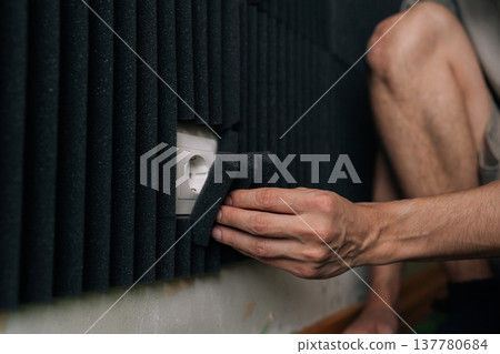 Closeup hand of professional hand attaching black acoustic foam panels to wall, working around electrical socket, transforming room into soundproof studio space for audio recording noise reduction. Closeup hand of professional hand attaching black acoustic foam panels to wall, working around electrical socket, transforming room into soundproof studio space for audio recording noise reduction. 137780684