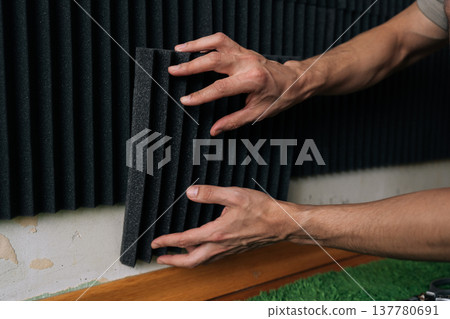 Person installing black acoustic absorbing foam panels onto a wall, creating a soundproof environment for a recording studio or home entertainment space. 137780691