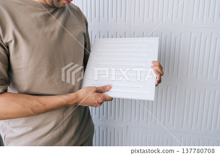 Man holding a square acoustic panel, preparing to install soundproofing material onto a textured white wall, focusing on noise reduction and enhancing room acoustics. Man holding a square acoustic panel, preparing to install soundproofing material onto a textured white wall, focusing on noise reduction and enhancing room acoustics. 137780708
