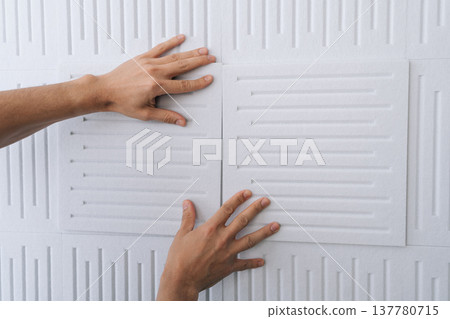 Close-up hand of handyman applying white soundproof acoustic panels onto a wall, improving home interior acoustics and soundproofing a room during renovation or construction. Close-up hand of handyman applying white soundproof acoustic panels onto a wall, improving home interior acoustics and soundproofing a room during renovation or construction. 137780715