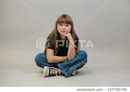 A young girl in a black t-shirt and wide-leg jeans sits casually on the floor with her hand resting on her knee against a neutral background. This image highlights a relaxed, casual fashion style for A young girl in a black t-shirt and wide-leg jeans sits casually on the floor with her hand resting on her knee against a neutral background. This image highlights a relaxed, casual fashion style for 137780746