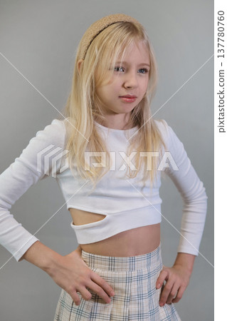 A young girl with blonde hair, wearing a white crop top and plaid skirt, poses confidently with her hands on her hips against a neutral background. This image highlights youthful fashion and a strong A young girl with blonde hair, wearing a white crop top and plaid skirt, poses confidently with her hands on her hips against a neutral background. This image highlights youthful fashion and a strong 137780760