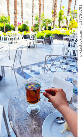 Close view of a person holding a cup of hot tea with lemon while sitting at a sunny caf terrace. The peaceful travel moment highlights slow living, morning relaxation and holiday lifestyle. Close view of a person holding a cup of hot tea with lemon while sitting at a sunny caf terrace. The peaceful travel moment highlights slow living, morning relaxation and holiday lifestyle. 137780791