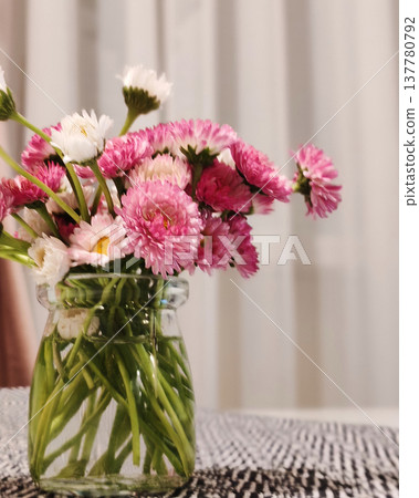 Delicate pink and white flowers arranged in a simple glass jar creating a charming tabletop decoration. The natural interior scene illustrates modern home decor, calm atmosphere and aesthetic living 137780792