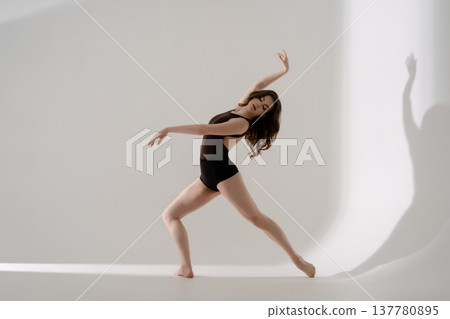 Expressive female dancer in a dark leotard performs a contemporary routine in a minimalist white studio. 137780895