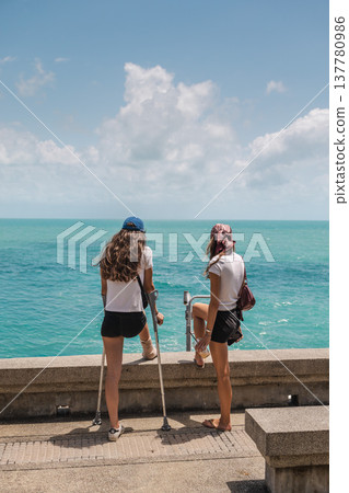 Two young women standing on seaside promenade looking at turquoise ocean 137780986