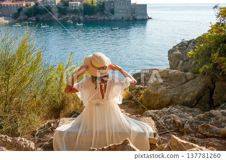 Woman in Hat and White Dress Sitting on Rocks with Sveti Stefan Island in Background 137781260