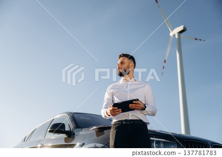 Standing by the car, holding digital tablet. Engineer in field checking on wind turbine production 137781783