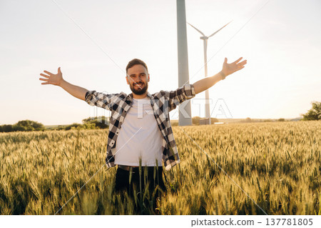 Hands to the sides, happy. Guy engineer in wheat field against wind turbines Hands to the sides, happy. Guy engineer in wheat field against wind turbines 137781805