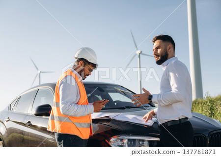 Black car, standing near it and talking. Two men engineers are on field, with wind turbines 137781871