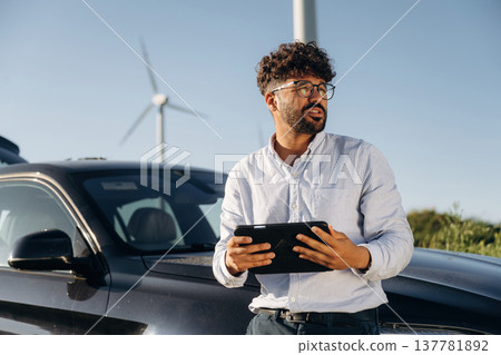 By the front of the car, with tablet. Male engineer in field checking on wind turbine production By the front of the car, with tablet. Male engineer in field checking on wind turbine production 137781892