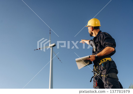 Pointing to the wind turbine by hand. Engineer in field on clean energy production Pointing to the wind turbine by hand. Engineer in field on clean energy production 137781979