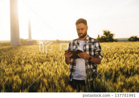 Sunny weather, front view. Guy engineer in wheat field against wind turbines Sunny weather, front view. Guy engineer in wheat field against wind turbines 137781991