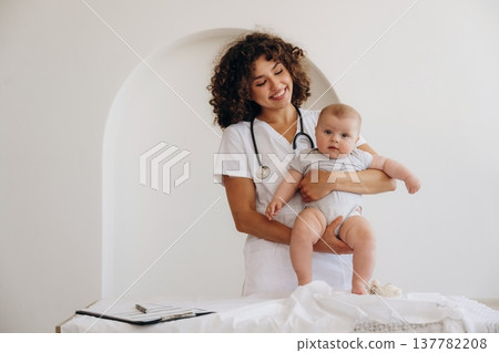 In coat, with stethoscope. Holding kid in hands. Female doctor is with newborn baby indoors In coat, with stethoscope. Holding kid in hands. Female doctor is with newborn baby indoors 137782208