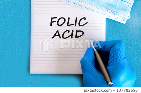Folic acid symbol. Concept words Folic acid on beautiful white note. Doctor's hand in blue medical glove. Medical mask. Beautiful blue background. Medical healthy lifestyle folic acid concept. Folic acid symbol. Concept words Folic acid on beautiful white note. Doctor's hand in blue medical glove. Medical mask. Beautiful blue background. Medical healthy lifestyle folic acid concept. 137782656