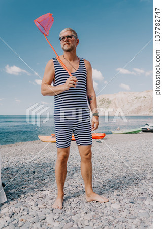 Man beach swimsuit standing on a pebble shore with a butterfly net during a sunny summer vacation by the sea 137782747