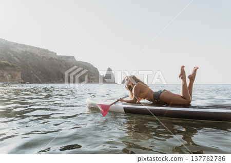 Woman Paddleboarding Relaxing Sea Coastline - A young woman lays on her paddleboard and relaxes in the sea while gazing at a coastal scene with mountains and cliffs in the background. 137782786