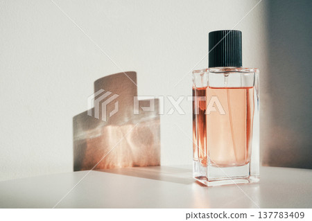 Glass perfume bottle standing on white surface with harsh sunlight creating shadow 137783409