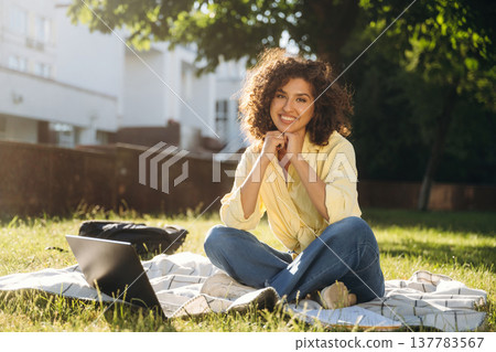 Looking forward, sitting on the blanket with laptop. Beautiful woman student is near the university outdoors Looking forward, sitting on the blanket with laptop. Beautiful woman student is near the university outdoors 137783567