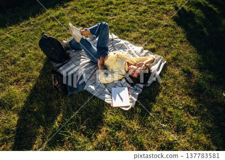 Happy woman student is laying down on a blanket on the grass Happy woman student is laying down on a blanket on the grass 137783581