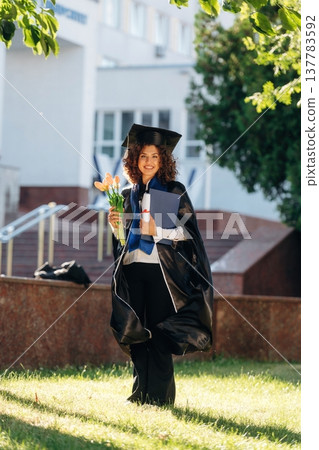 Green tree and university building behind. Cheerful graduate student in a gown is standing outdoors Green tree and university building behind. Cheerful graduate student in a gown is standing outdoors 137783592