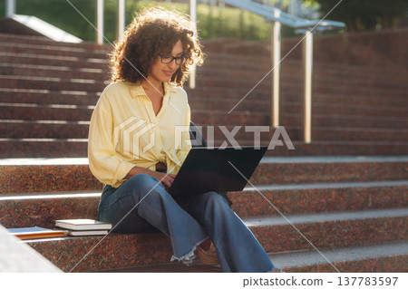 Female student is sitting on the stairs with laptop on her legs Female student is sitting on the stairs with laptop on her legs 137783597