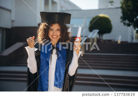 Diploma in hand. Cheerful graduate student in a gown is standing outdoors Diploma in hand. Cheerful graduate student in a gown is standing outdoors 137783619
