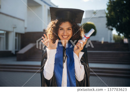 Finally finished, diploma in hand. Cheerful graduate student in a gown is standing outdoors Finally finished, diploma in hand. Cheerful graduate student in a gown is standing outdoors 137783623