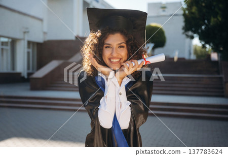 Finally finished, diploma in hand. Cheerful graduate student in a gown is standing outdoors Finally finished, diploma in hand. Cheerful graduate student in a gown is standing outdoors 137783624