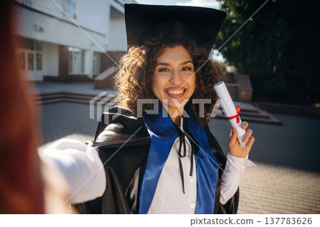Making a selfie, holding diploma document. Cheerful graduate student in a gown is standing outdoors 137783626