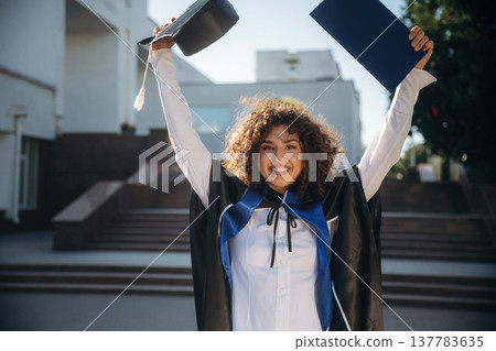 Joyful, with diploma. Cheerful graduate student in a gown is standing outdoors Joyful, with diploma. Cheerful graduate student in a gown is standing outdoors 137783635