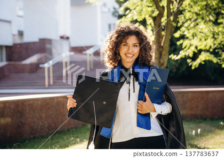 In official formal clothes, with diploma. Cheerful graduate student in a gown is standing outdoors In official formal clothes, with diploma. Cheerful graduate student in a gown is standing outdoors 137783637