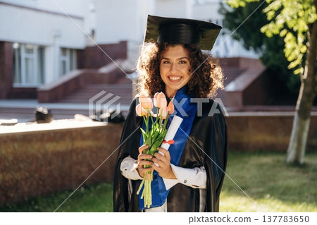Gift of flowers in hands. Cheerful graduate student in a gown is standing outdoors Gift of flowers in hands. Cheerful graduate student in a gown is standing outdoors 137783650